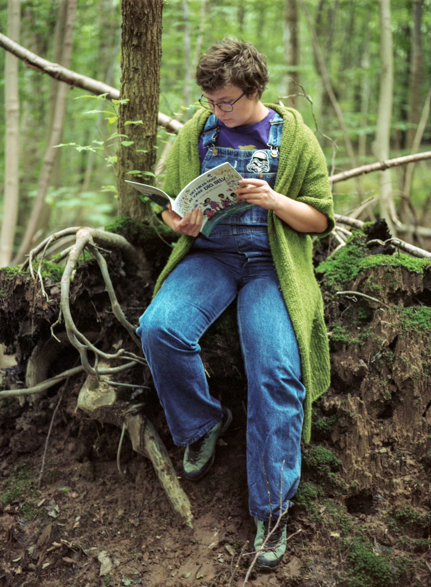 Udendørs foto af Ivy-Oak, en hvid nonbinær person, i en skov. De har kort brunt hår og blå briller på, de er iført denim overalls, en lang grøn strikket gardigan, lilla t-shirt og blå sko. De sidder på en lille forhøjning med den ene fod oppe på nogle trærødder og læser i billedbogen Det føles godt at være dig selv. Photo Credit: Astrid Ubaghs